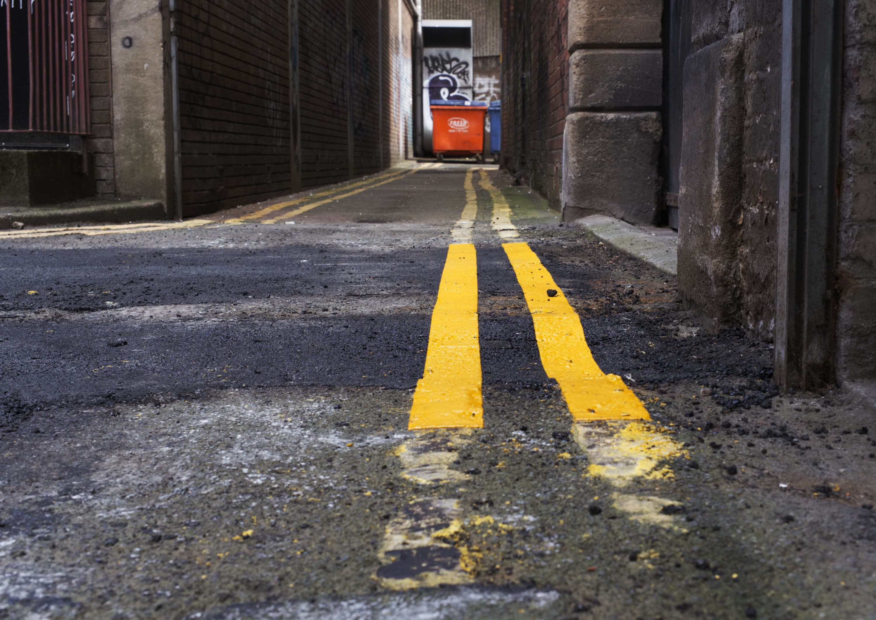 An almost ground level shot of yellow parking lines leading down an alley. The alley ends with a large orange bin.